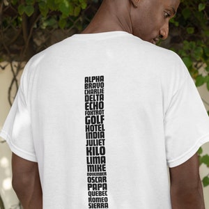 以下が含まれることがあります： NATOの音声アルファベットを黒文字で記した白いTシャツ。テキストは次のとおりです。アルファ、ブラボー、チャーリー、デルタ、エコー、フォックストロット、ゴルフ、ホテル、インディア、ジュリエット、キロ、リマ、マイク、ノベンバー、オスカー、パパ、キューベック、ロメオ、シエラ、タンゴ、ユニフォーム、ビクター、ウィスキー、エックスレイ、ヤンキー、ズールー。