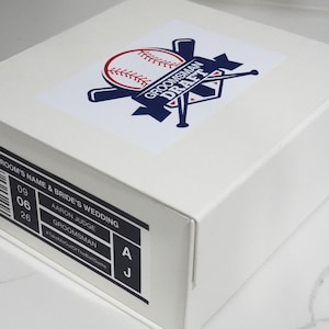 May include: A white box with a baseball-themed design. The design includes a baseball, bats, and the text "GROOMSMAN DRAFT." The box also features a label with wedding details, including the names and date.