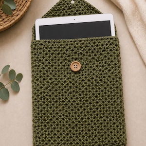 Op de afbeelding: Olijfgroene gehaakte tablethoes met een houten knoopsluiting. De hoes heeft een klepsluiting en een gestructureerd steekpatroon. Een witte tablet is gedeeltelijk zichtbaar. De hoes is ongeveer 25 cm hoog.