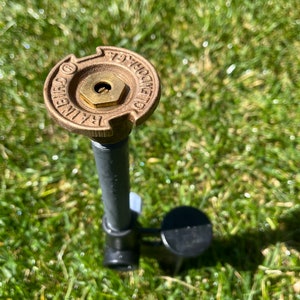 May include: A close-up of a brass sprinkler head with the text "1/2" and "GENUINE" on it. The sprinkler head is attached to a gray pipe and is sitting in green grass.