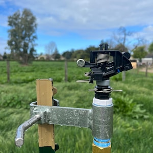 May include: A black sprinkler head attached to a metal pipe with a silver bracket. The sprinkler head is mounted on a green metal post. The sprinkler head is in a field of green grass.