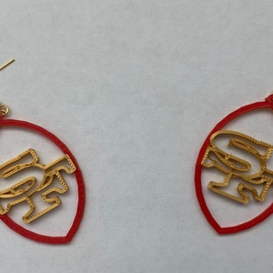 May include: A pair of red and gold earrings shaped like a football with the letters "SF" inside.
