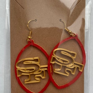 May include: A pair of red hoop earrings with gold-colored "SF" lettering. The earrings are packaged on a brown card.