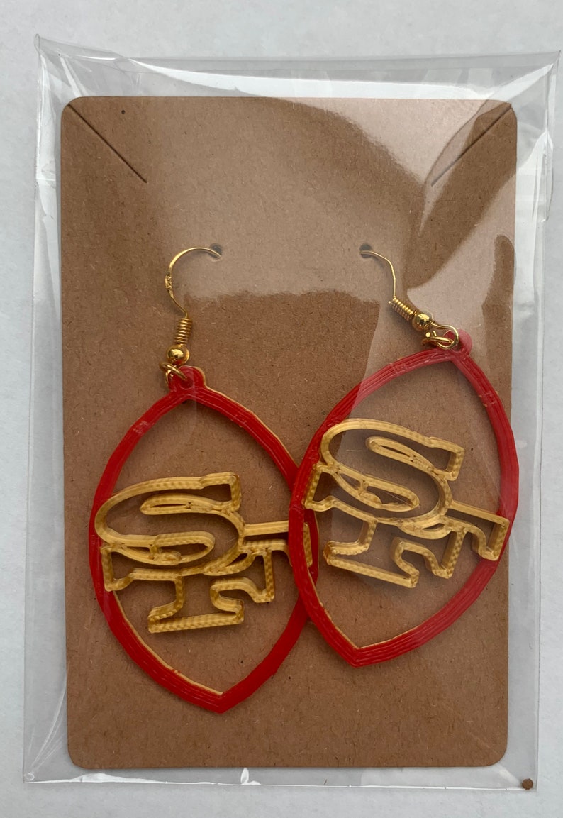 May include: A pair of red hoop earrings with gold-colored "SF" lettering. The earrings are packaged on a brown card.