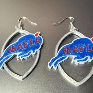 May include: A pair of earrings featuring a blue buffalo with the word "MAFIA" in red lettering. The buffalo is set against a white outline and a silver teardrop-shaped frame. The earrings have silver hooks.
