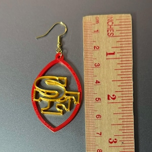 May include: Red and gold football-shaped earrings with the letters "SF" inside. The earrings are about 2 inches long.