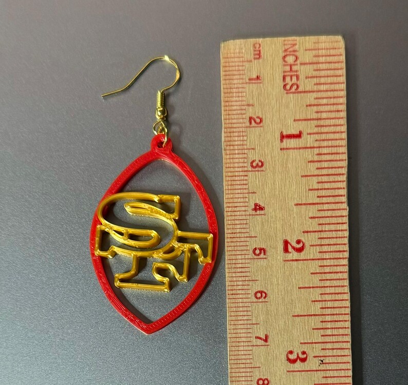 May include: Red and gold football-shaped earrings with the letters "SF" inside. The earrings are about 2 inches long.
