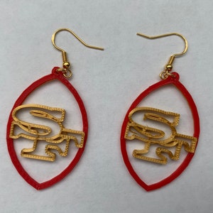May include: A pair of red and gold earrings shaped like a football with the letters "SF" inside. The earrings are made of plastic and have gold hooks.