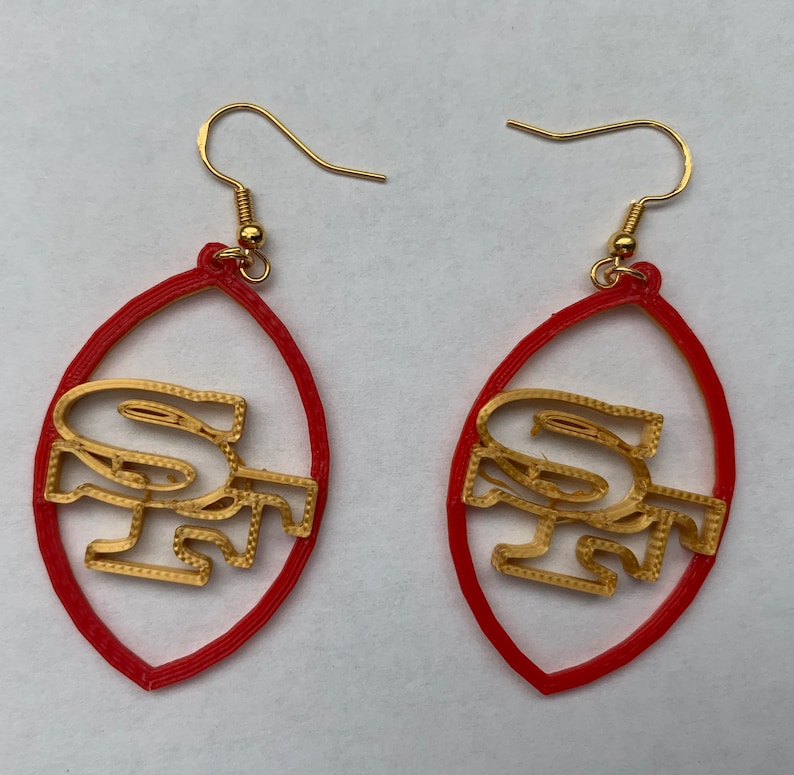 May include: A pair of red and gold earrings shaped like a football with the letters "SF" inside. The earrings are made of plastic and have gold hooks.