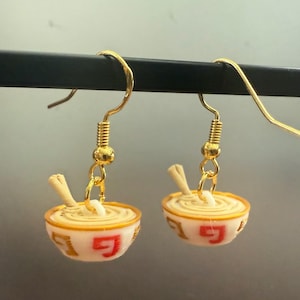 May include: A pair of gold dangle earrings featuring miniature bowls of ramen noodles. The bowls are white with red and gold accents. The noodles are white and the chopsticks are brown.