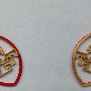 May include: A pair of gold-colored earrings shaped like a football with the letters "SF" inside. The earrings are red on the outside and gold on the inside.