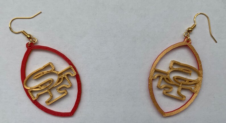 May include: A pair of gold-colored earrings shaped like a football with the letters "SF" inside. The earrings are red on the outside and gold on the inside.