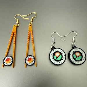 May include: Sushi-themed earrings. One pair features gold-colored chopsticks with a sushi roll at the base. The other pair has a sushi roll design with a black border and silver-colored hooks. The sushi rolls have orange, green, and white details.