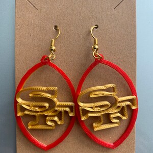 May include: A pair of red and gold earrings shaped like a football with the San Francisco 49ers logo.