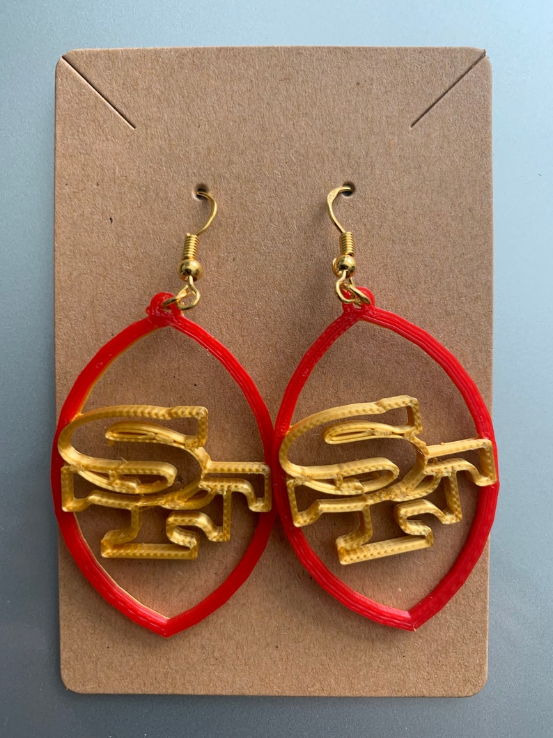 May include: A pair of red and gold earrings shaped like a football with the San Francisco 49ers logo.