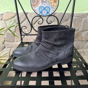 May include: A pair of charcoal gray leather ankle boots featuring a rounded toe, a low block heel, and a soft, ruched texture across the vamp. The boots have a simple slip-on design with a clean, unadorned shaft and a matching dark rubber outsole.
