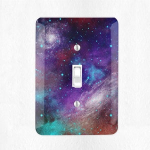 May include: A light switch cover with a galaxy design. The cover features a dark blue, purple, and green nebula with a scattering of white stars.
