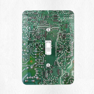 May include: A light switch cover with a green circuit board design. The switch is white and is in the center of the cover.