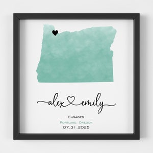 May include: Framed art print featuring a teal watercolor map of Oregon with a black heart marking a location. The names "alex & emily" are written in cursive, with the word "engaged" and the date 07.31.2025 below.