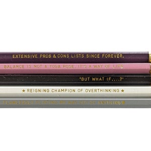 May include: Set of six pencils with different colored wood and gold lettering. The pencils have the following sayings: "Extensive pros & cons lists since forever", "Balance is not a yoga pose, it's a way of life", "'But what if...?'", "★Reigning champion of overthinking★", "Flawlessly talking my way out of anything".