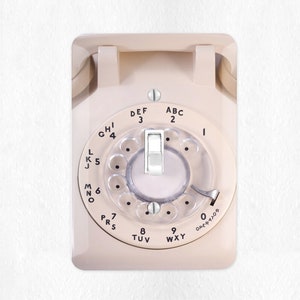 May include: A vintage rotary phone with a white face and black lettering. The phone has a rotary dial with numbers 1 through 0 and letters ABC, DEF, GHI, JKL, MNO, PRS, TUV, WXY. The phone is mounted on a white wall.