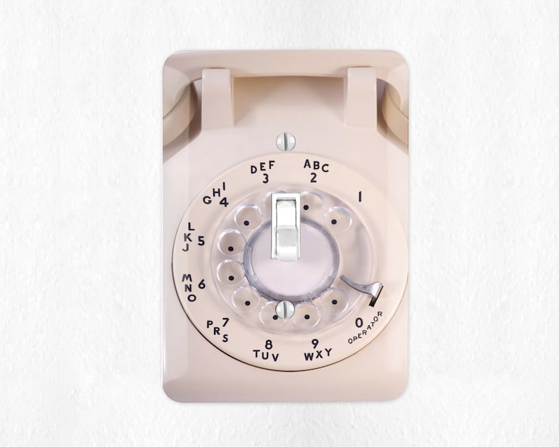 May include: A vintage rotary phone with a white face and black lettering. The phone has a rotary dial with numbers 1 through 0 and letters ABC, DEF, GHI, JKL, MNO, PRS, TUV, WXY. The phone is mounted on a white wall.