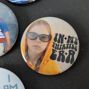 May include: A round button with a photo of a young girl wearing glasses and a yellow hoodie. The text "IN MY THIRTIES ERA" is printed in black with a retro font.