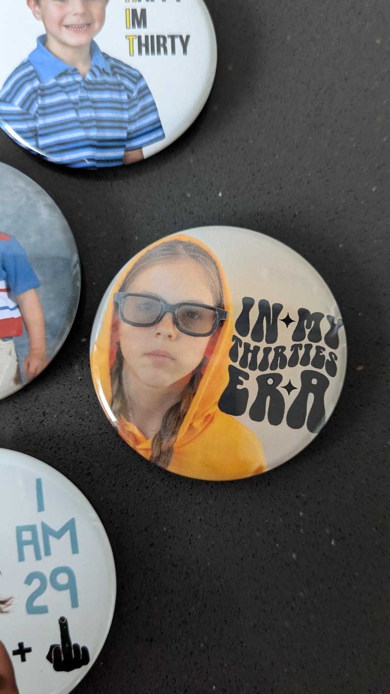 May include: A round button with a photo of a young girl wearing glasses and a yellow hoodie. The text "IN MY THIRTIES ERA" is printed in black with a retro font.