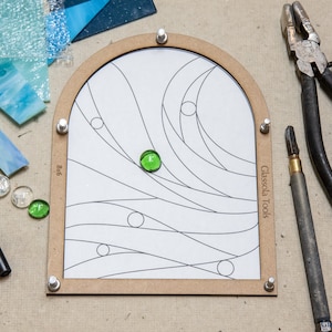 Peut inclure: Un projet de vitrail en cours sur un cadre en bois avec un sommet arrondi. Le cadre présente un motif de lignes et de cercles dessinés. De petits morceaux de verre vert sont placés sur le cadre. Le cadre est étiqueté "Glassola Tools".