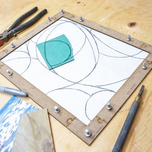 May include: A stained glass project in progress. A wooden frame holds a piece of glass with a design drawn on it. A piece of green glass is placed on top of the design. A silver marker, a glass cutter, and a pair of pliers are also visible.