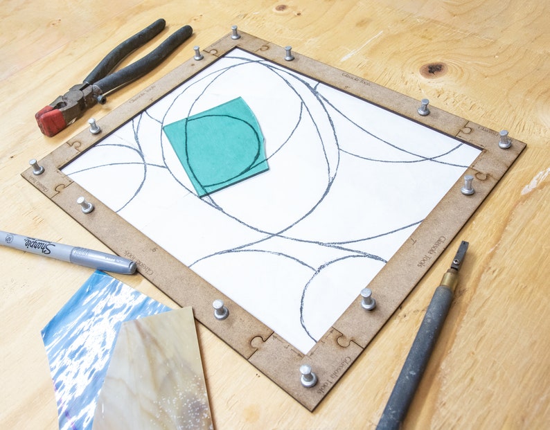 May include: A stained glass project in progress. A wooden frame holds a piece of glass with a design drawn on it. A piece of green glass is placed on top of the design. A silver marker, a glass cutter, and a pair of pliers are also visible.