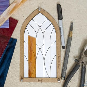 May include: A stained glass pattern template with a brown wooden frame. The template is 8x4 inches and has a design of a pointed arch window with a decorative pattern of curved lines. A piece of clear glass is placed on the template. The template is on a beige surface with a black Sharpie marker, a metal tool, and a pair of pliers.