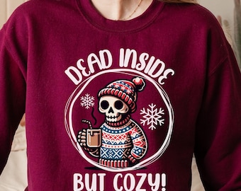 Puede incluir: Una sudadera color burdeos con un gráfico blanco de un esqueleto con un gorro de invierno y sosteniendo una taza. El texto "Dead Inside But Cozy!" está impreso en la sudadera.