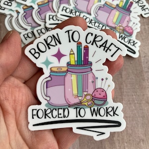 Puede incluir: Pegatina blanca con el texto "Born to Craft Forced to Work" en negro. La pegatina presenta una ilustración de dibujos animados de dos tarros de cristal llenos de lápices, tijeras y un alfiletero.