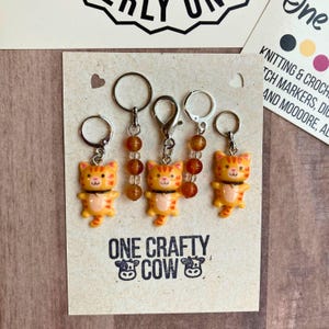 May include: Set of three orange and white cat stitch markers with amber beads. The markers are attached to silver rings and have a silver clasp. The card reads "One Crafty Cow".