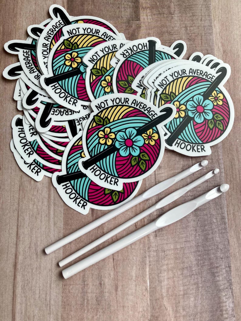 Puede incluir: Una pila de pegatinas redondas de colores con el texto "Not Your Average Hooker" y un gr&aacute;fico de un gancho de ganchillo y lana. Tambi&eacute;n se muestran tres ganchos de ganchillo blancos.