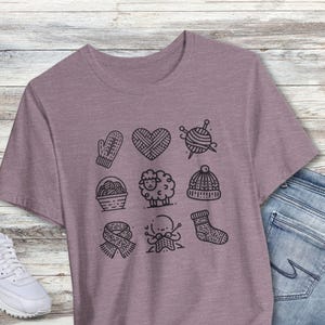 May include: A heather purple t-shirt with a graphic design of nine knitting-themed icons. The icons include a mitten, a heart, yarn, a needle, a sheep, a basket, a scarf, a hand knitting, and a sock.