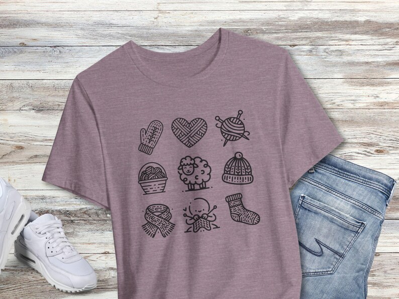 May include: A heather purple t-shirt with a graphic design of nine knitting-themed icons. The icons include a mitten, a heart, yarn, a needle, a sheep, a basket, a scarf, a hand knitting, and a sock.