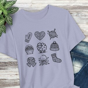 May include: A lavender t-shirt with a graphic design of various knitting-related icons, including a heart, a ball of yarn, a pair of knitting needles, a mitten, a basket of yarn, a sheep, a hat, a pair of knitting needles, and a sock.