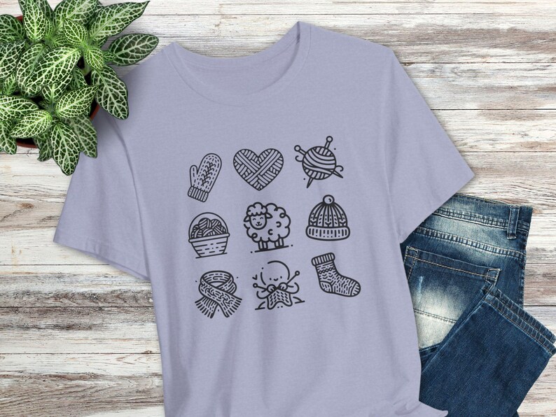 May include: A lavender t-shirt with a graphic design of various knitting-related icons, including a heart, a ball of yarn, a pair of knitting needles, a mitten, a basket of yarn, a sheep, a hat, a pair of knitting needles, and a sock.