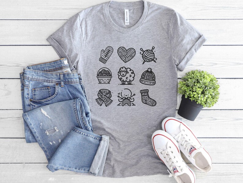 A medium grey tee shirt with 9 knitting icons in rows including a sheep, a hat, a sock, a basket of yarn, a mitten, a heart shape, a ball of yarn with knitting needles, a scarf, and a figure knitting