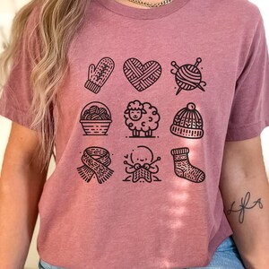 May include: A mauve t-shirt with a graphic of nine black and white illustrations of knitting-related items, including a mitten, a heart, yarn balls, a sheep, a hat, a scarf, and a sock.