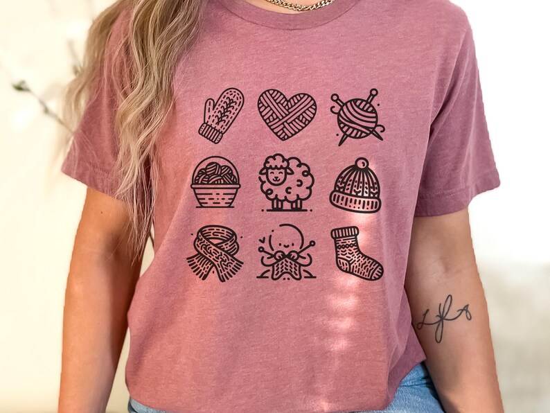 May include: A mauve t-shirt with a graphic of nine black and white illustrations of knitting-related items, including a mitten, a heart, yarn balls, a sheep, a hat, a scarf, and a sock.
