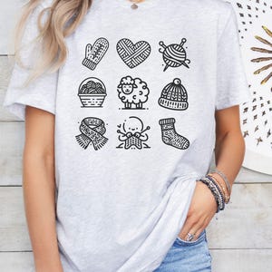 May include: A gray t-shirt with a black and white graphic design of nine knitting-themed icons. The icons include a mitten, a heart, yarn, a sheep, a hat, a scarf, a pair of knitting needles, and a sock.