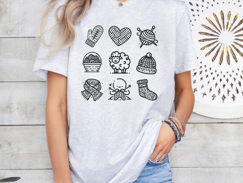 May include: A gray t-shirt with a black and white graphic design of nine knitting-themed icons. The icons include a mitten, a heart, yarn, a sheep, a hat, a scarf, a pair of knitting needles, and a sock.