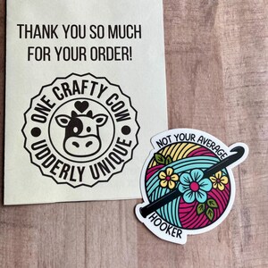 Puede incluir: Tarjeta de agradecimiento con una ilustraci&oacute;n de una vaca en blanco y negro y el texto "One Crafty Cow Uddderly Unique". Tambi&eacute;n se muestra una pegatina con una bola de hilo de colores, un gancho de ganchillo y el texto "Not Your Average Hooker".