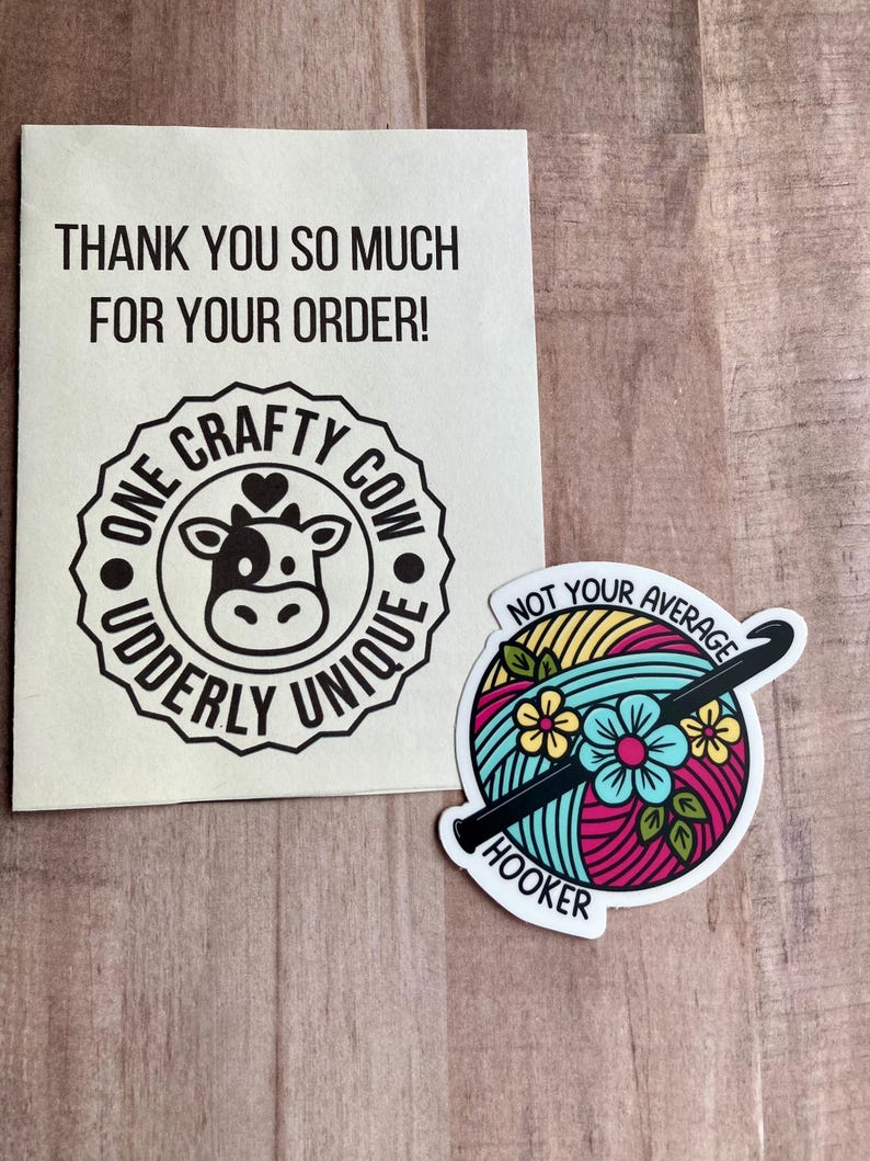 Puede incluir: Tarjeta de agradecimiento con una ilustraci&oacute;n de una vaca en blanco y negro y el texto "One Crafty Cow Uddderly Unique". Tambi&eacute;n se muestra una pegatina con una bola de hilo de colores, un gancho de ganchillo y el texto "Not Your Average Hooker".