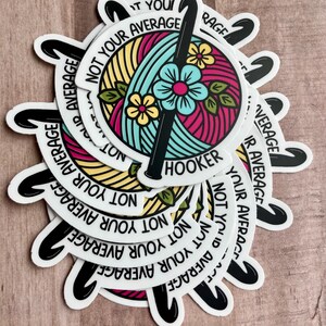Puede incluir: Una pila de pegatinas redondas con un dise&ntilde;o de gancho de ganchillo y ovillo de lana. Las pegatinas son blancas con texto negro que dice "Not Your Average Hooker".