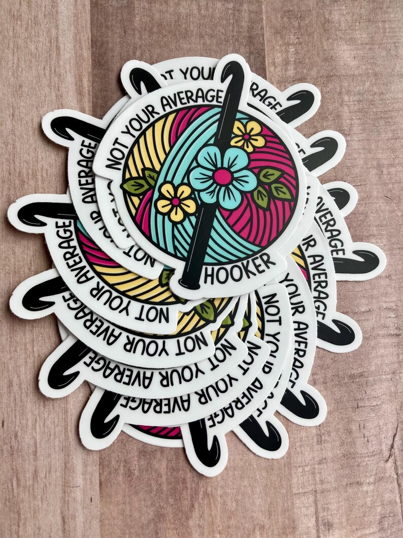 Puede incluir: Una pila de pegatinas redondas con un dise&ntilde;o de gancho de ganchillo y ovillo de lana. Las pegatinas son blancas con texto negro que dice "Not Your Average Hooker".