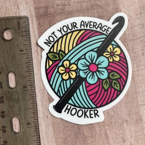 Puede incluir: Una pegatina blanca con una bola de hilo de colores y un gancho de ganchillo negro. El texto en la pegatina dice "Not Your Average Hooker".